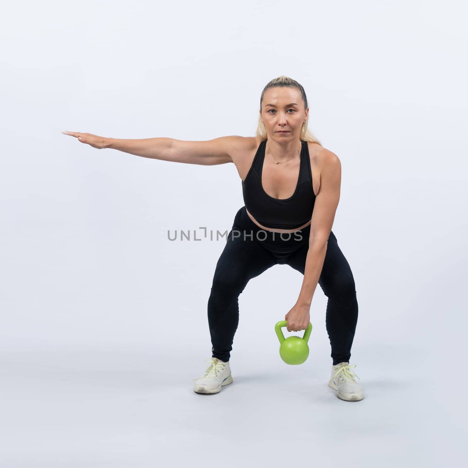 Full body length shot athletic and sporty senior woman doing squat with kettlebell for body workout on isolated background. Healthy active physique and body care lifestyle after retirement. Clout
