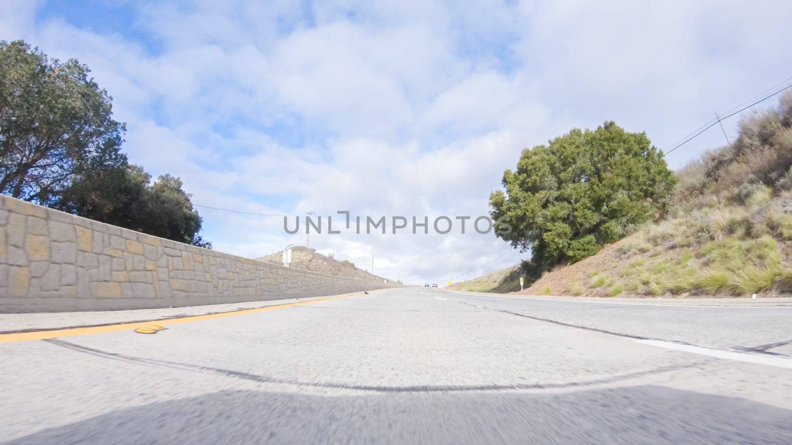 On a crisp winter day, a car cruises along the iconic Highway 101 near San Luis Obispo, California. The surrounding landscape is brownish and subdued, with rolling hills and patches of coastal vegetation flanking the winding road.