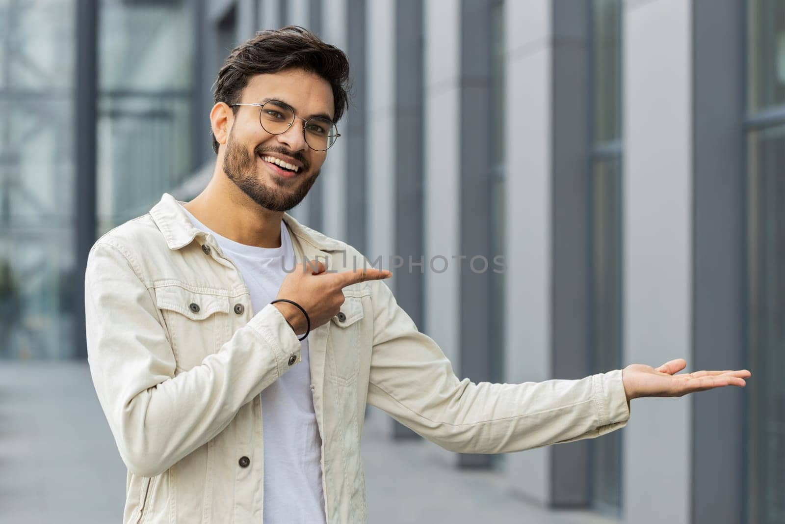 Happy Indian man showing pointing empty place advertising area for commercial text copy-space for goods promotion sale discount outdoors. Arabian young guy in downtown city street. Business people