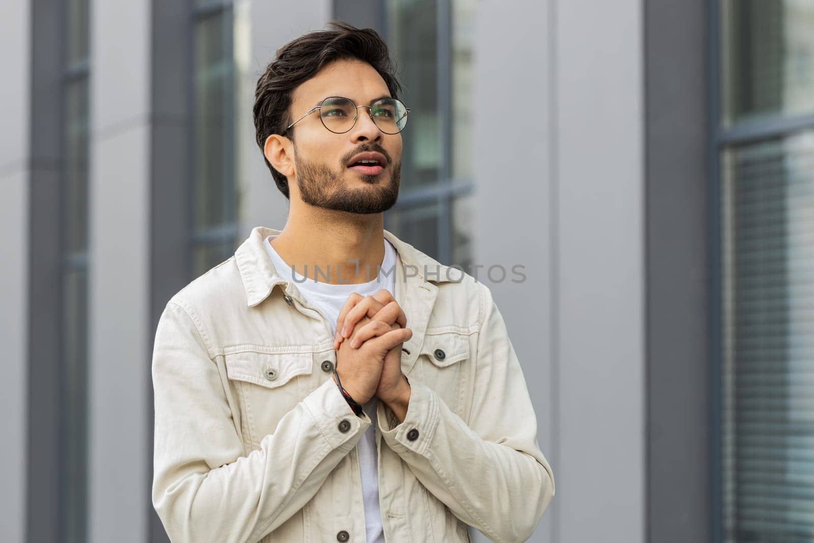 Young Indian man praying with closed eyes to God asking for blessing, help, forgiveness outdoors. Arabian Hindu guy clasping hands wishing luck in downtown city street. Business people lifestyles
