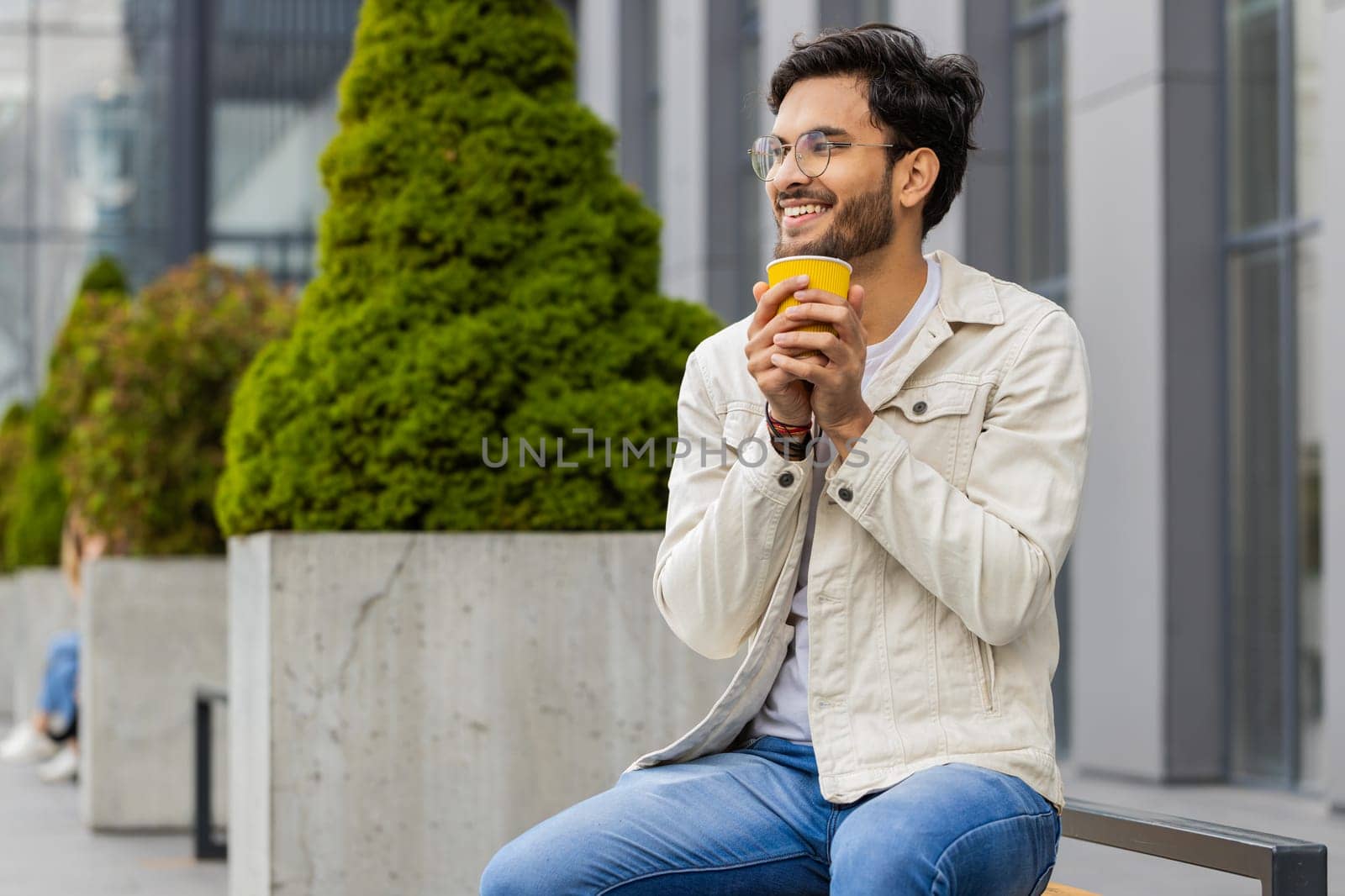 Happy Indian man enjoying morning coffee hot drink and smiling outdoors. Relaxing, taking a break. Arabian guy in city downtown street sitting on bench, drinking coffee to go. Town lifestyles outside