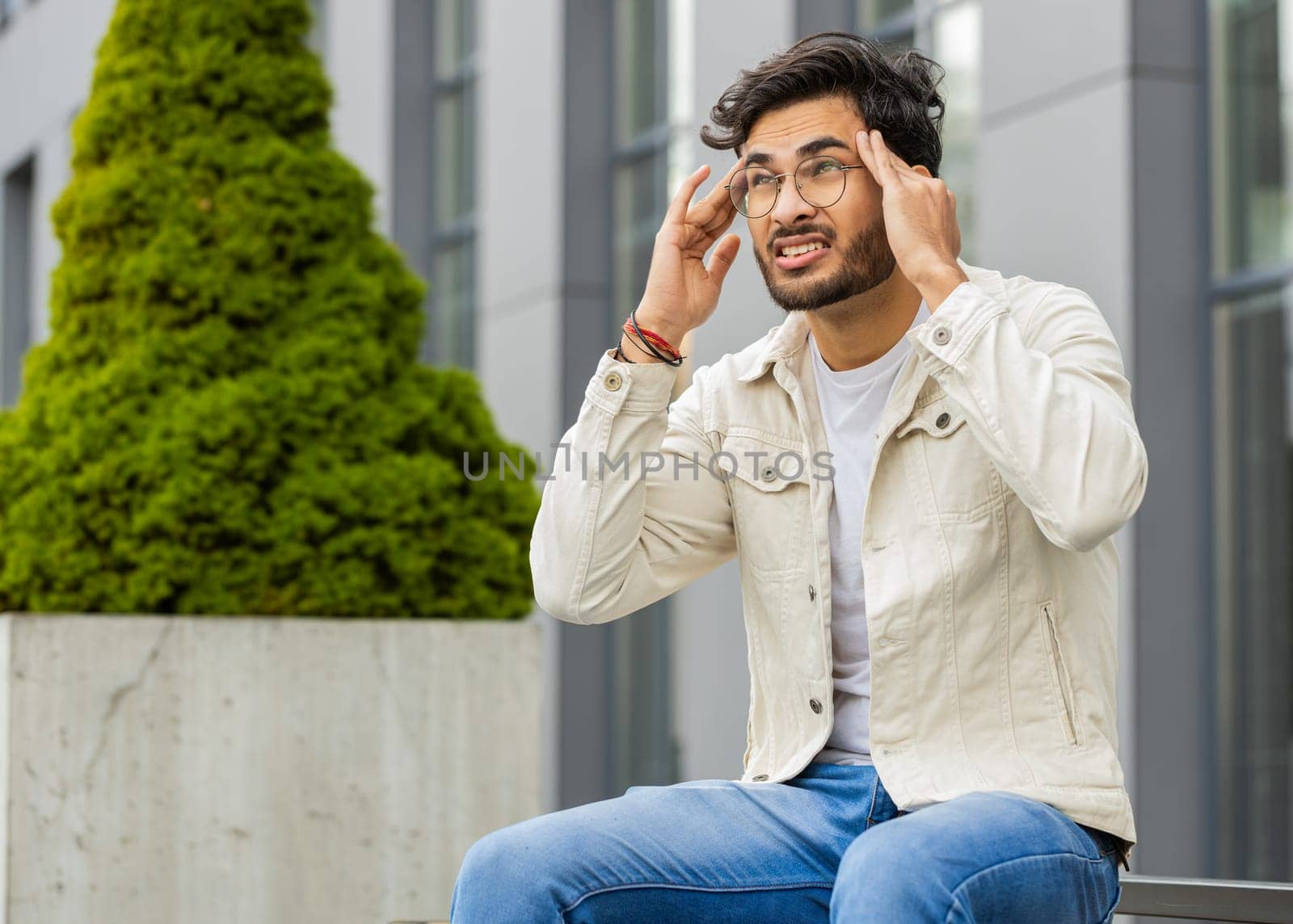 Displeased sick ill Indian man rubbing temples to cure headache problem, suffering from tension migraine, stress, grimacing in pain, high blood pressure outdoors. Arabian guy sitting on street bench