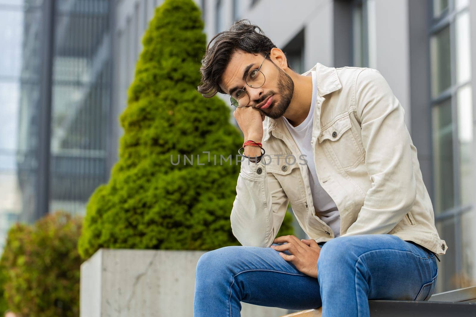 Tired sad Indian man feeling worried about financial problem stress outdoors. Arabian Hindu guy frustrated thinking of money debt budget loss bankruptcy sitting on downtown street bench. Overworking