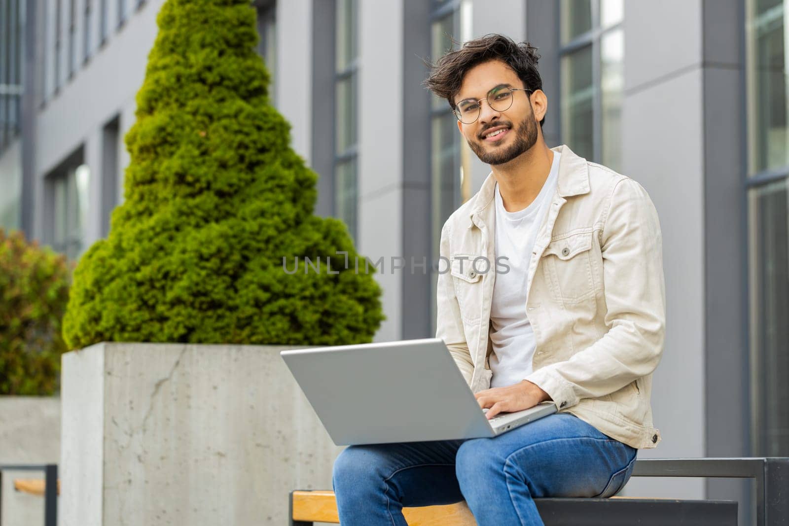 Happy Indian man freelancer working on laptop computer sends messages, makes online purchases, watching movies, reading email outdoors. Arabian Hindu guy looking at camera sitting on street bench
