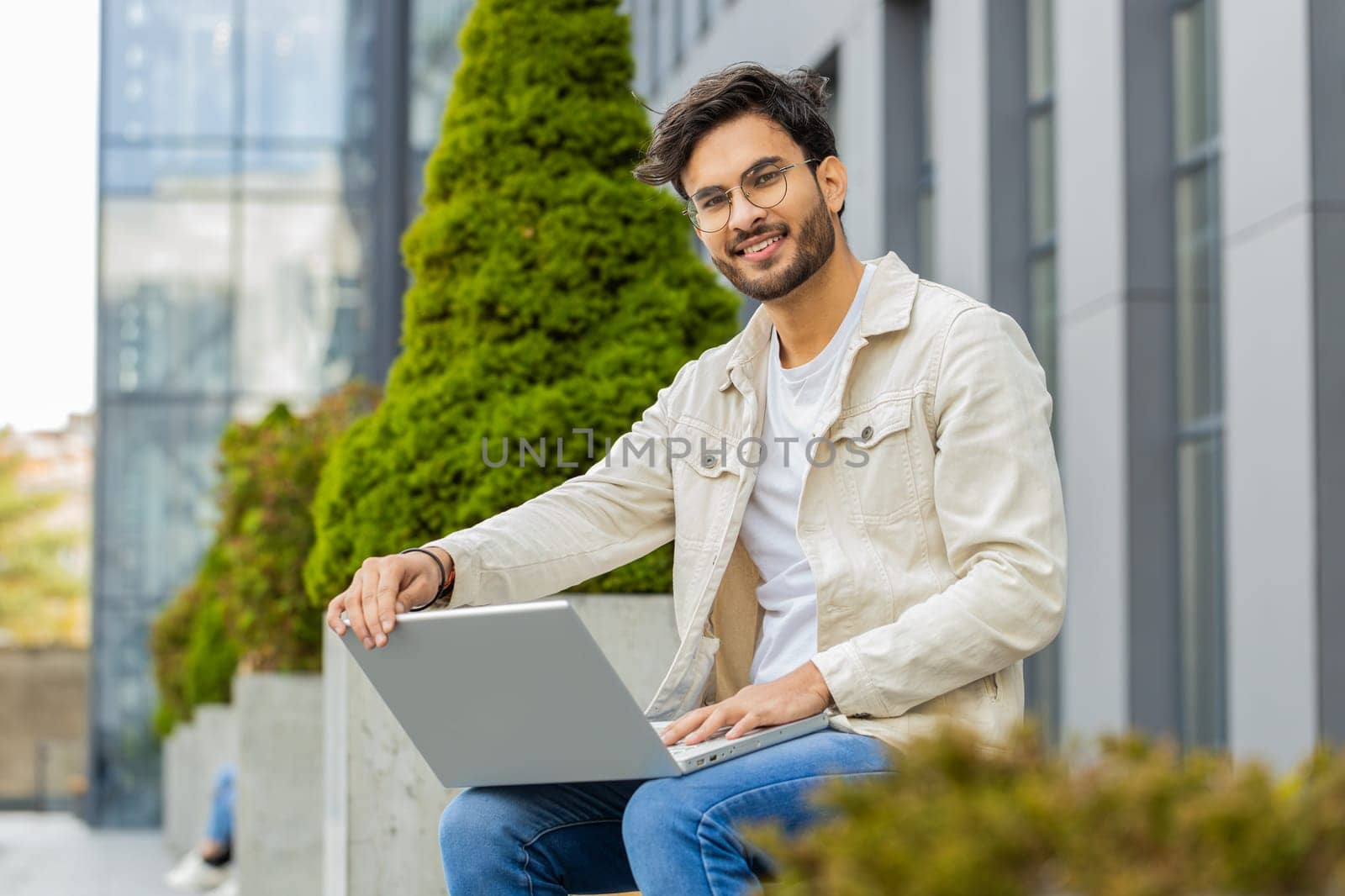 Happy young Indian man freelancer start working opens laptop sends messages, makes online purchases, watching movies, reading email outdoors. Arabian Hindu guy sitting on street bench. Business people