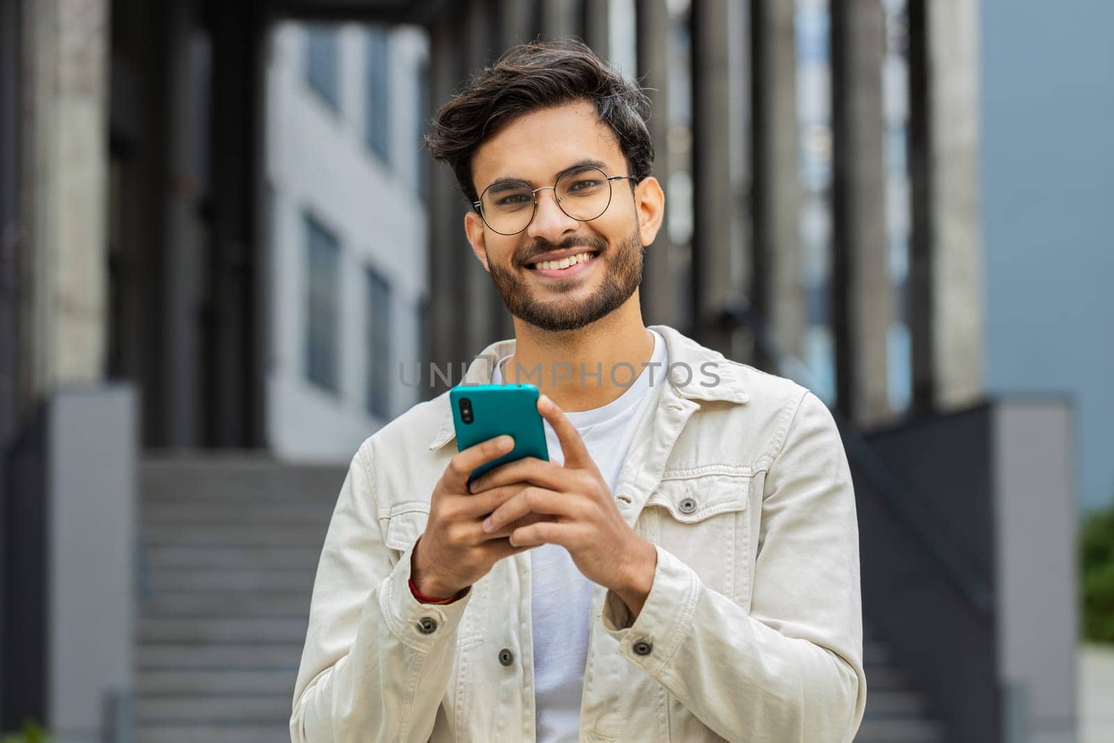 Young smiling Indian man using smartphone typing text messages, reading email, browsing news outdoors. Arabian Hindu guy use mobile social media app looking at camera standing in downtown city street