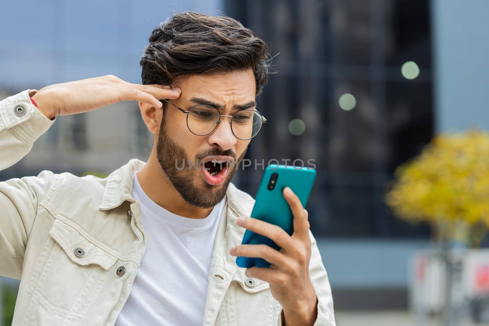 Displeased Indian man use smartphone typing browsing, loses becoming surprised sudden lottery results, bad news, fortune loss, fail outdoors. Arabian Hindu guy in downtown city street. Business people