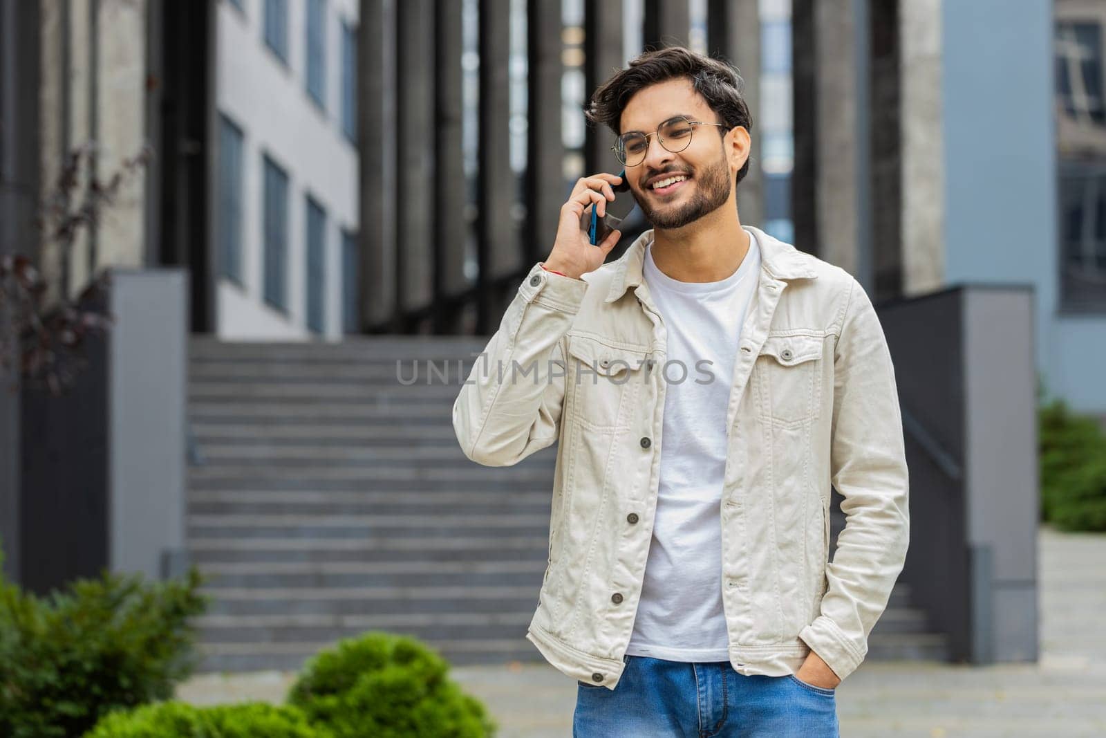 Young Indian man having remote conversation communicate speaking by smartphone outdoors. Arabian Hindu guy talking on phone unexpected good news gossip walking in downtown city street. Business people