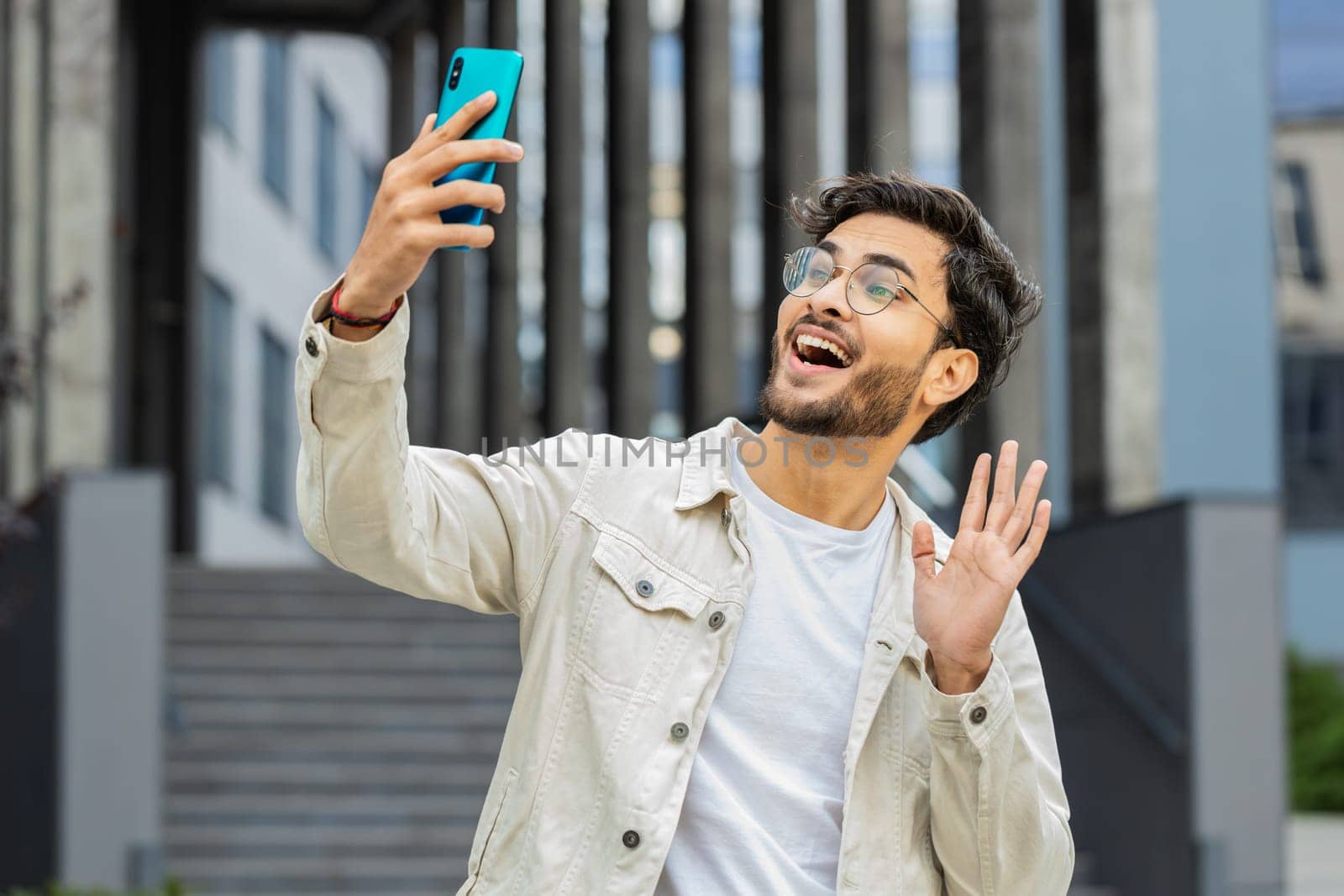 Young Indian man blogger taking selfie on smartphone, communicating video call online with subscribers recording stories for social media vlog outdoors. Arabian Hindu guy standing in urban city street