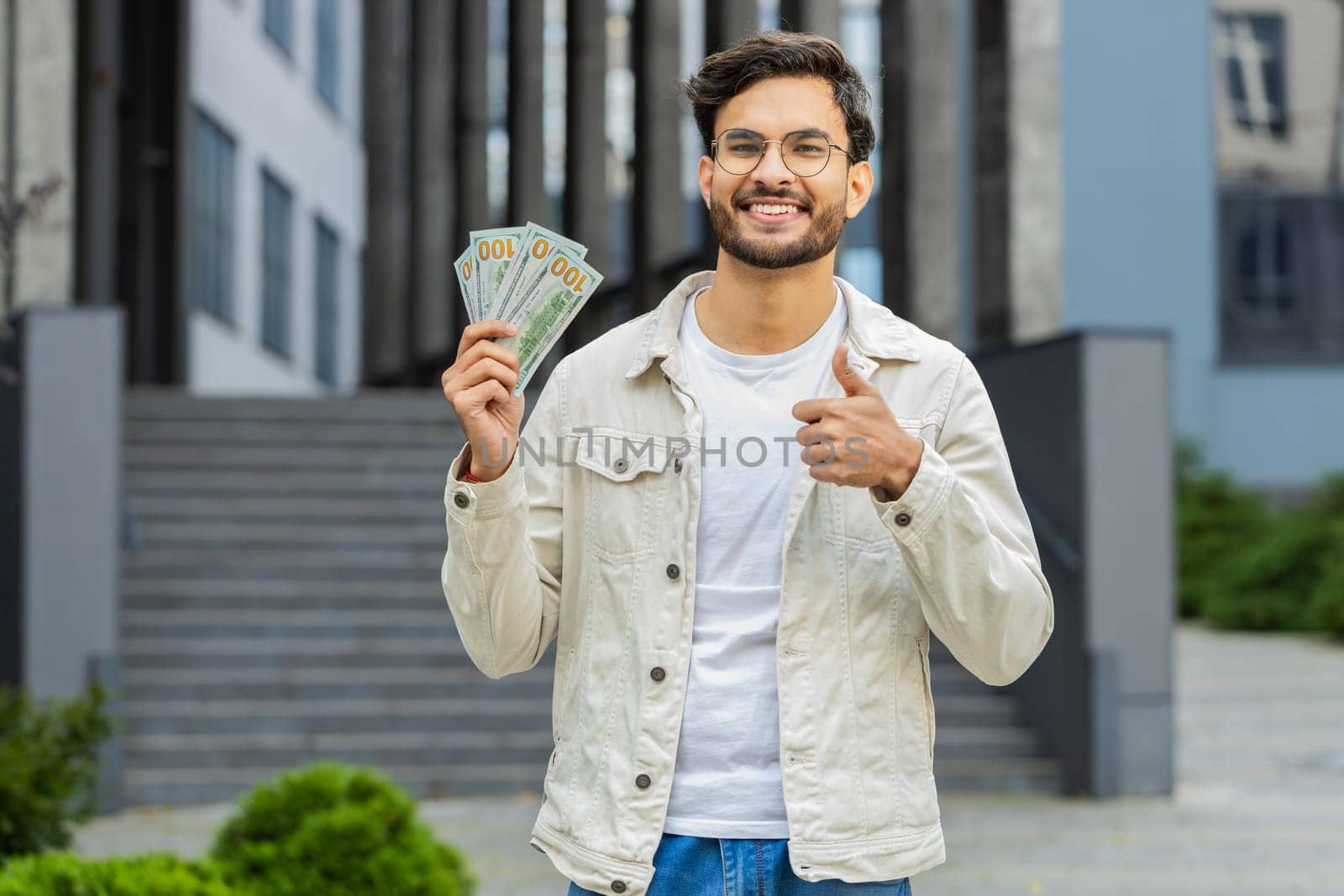 Happy Indian man counting money cash holding dollars banknotes, bills budget count salary profit reward bribe outdoors. Successful rich Arabian Hindu guy showing thumbs up in downtown city street