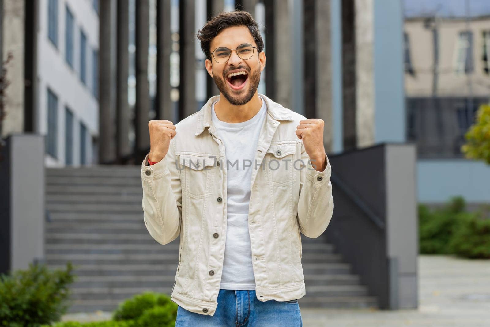 Happy Indian man entrepreneur shouting, celebrating success, winning, goal achievement, good news, lottery jackpot luck, victory outdoors. Arabian Hindu guy in downtown city street. Business people