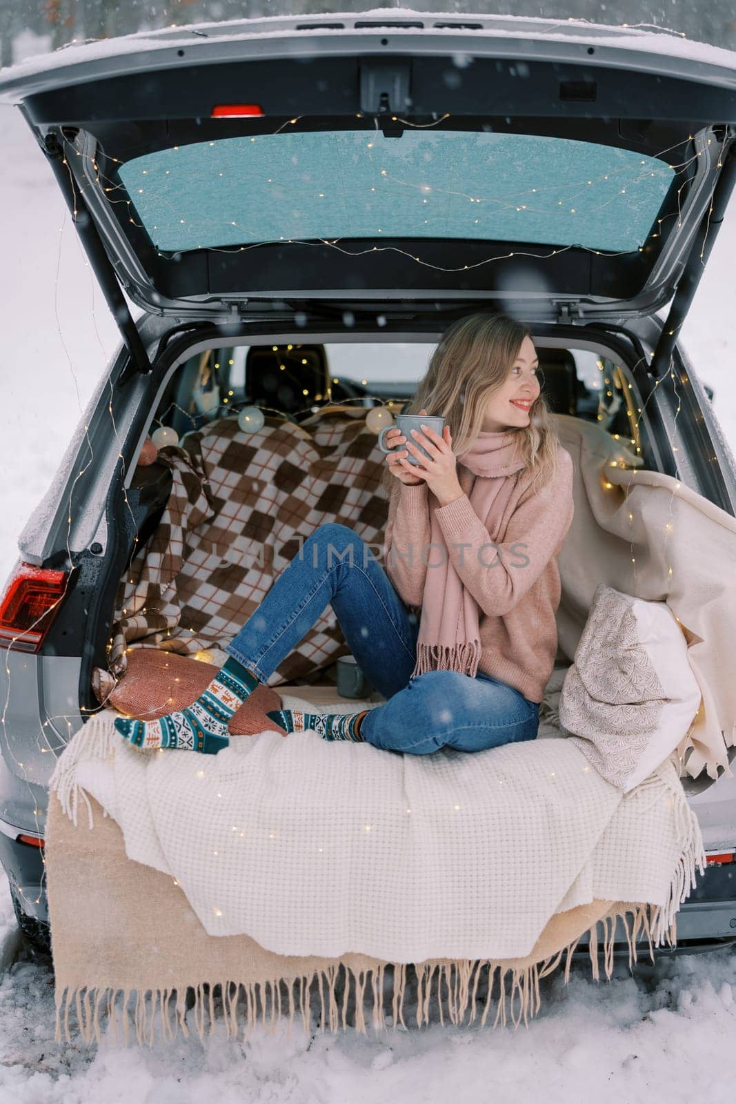 Girl with a mug of tea sits on a blanket in the trunk of a car in a snowy forest, turning her head to the side. High quality photo