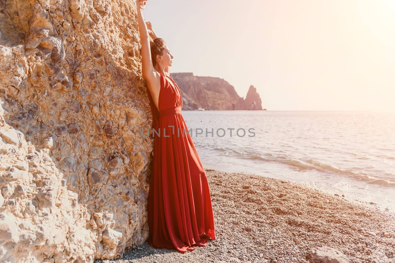 Woman in red dress on sea. Side view a Young beautiful sensual woman in a red long dress posing on the beach near sea on sunset. Girl on the nature on blue sky background. Fashion photo. by panophotograph