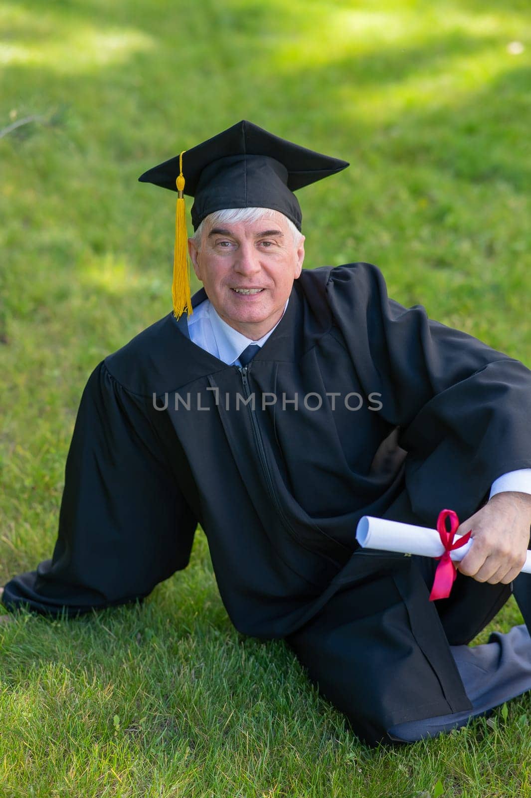 An elderly gray-haired man in a graduation gown sits on green grass. Vertical