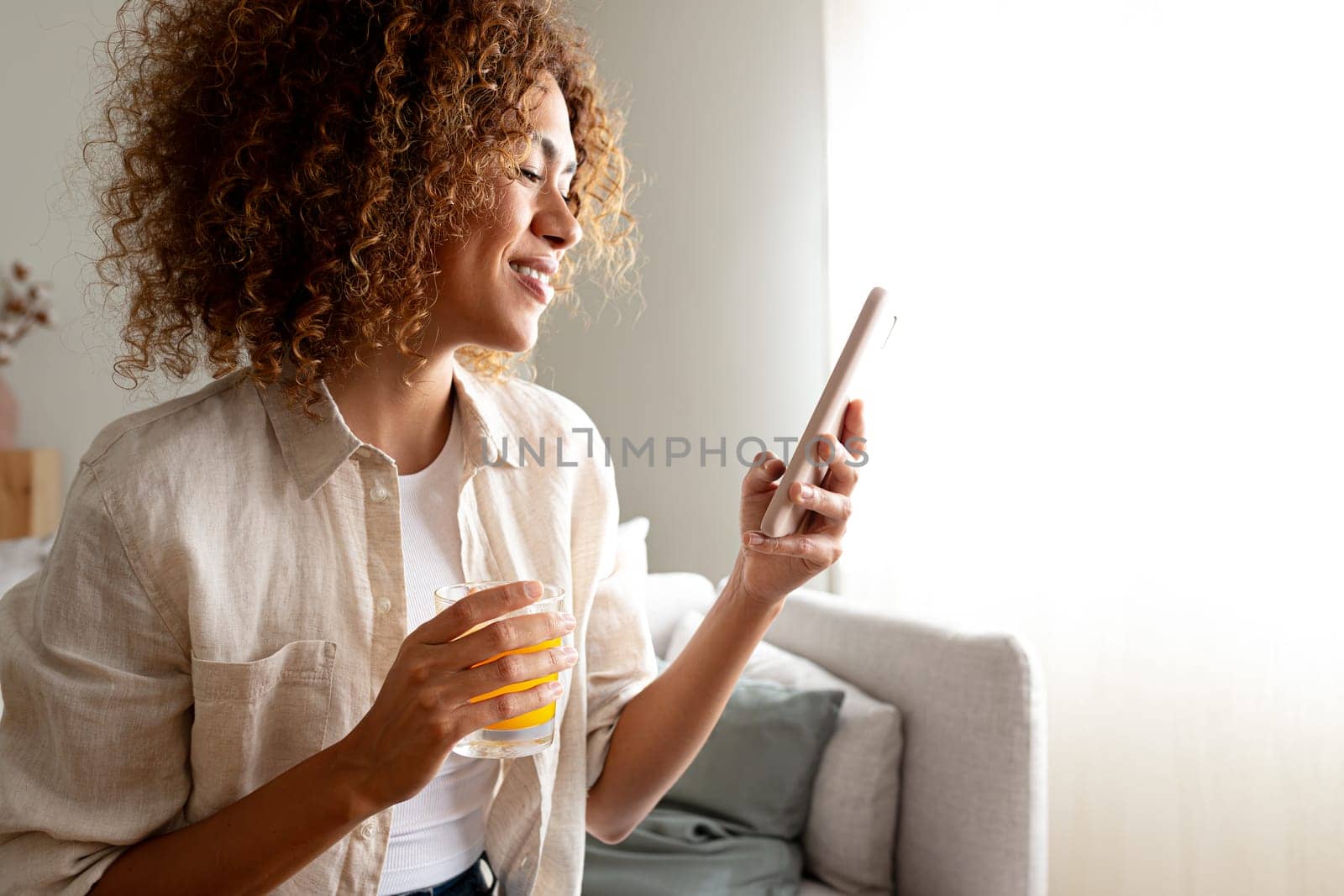 Happy woman using smart phone while drinking orange juice sitting on the couch at home cozy living room. Copy space. Lifestyle and technology concept.