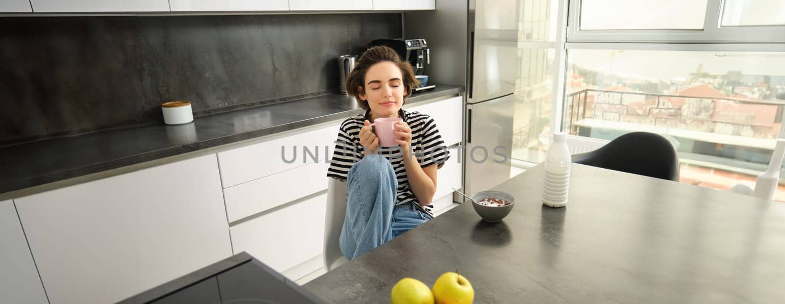 Young woman having breakfast or lunch, eating cereals with milk, drinking hot tea, sitting cosy in the kitchen.
