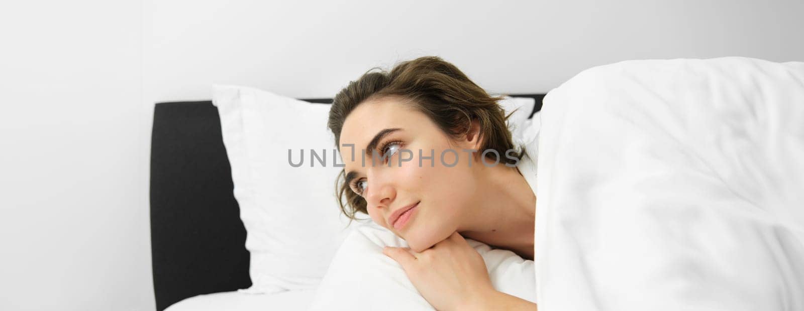 Close up portrait of beautiful brunette woman, waking up in her bed, looking at alarm clock, staring aside with smiling dreamy face.