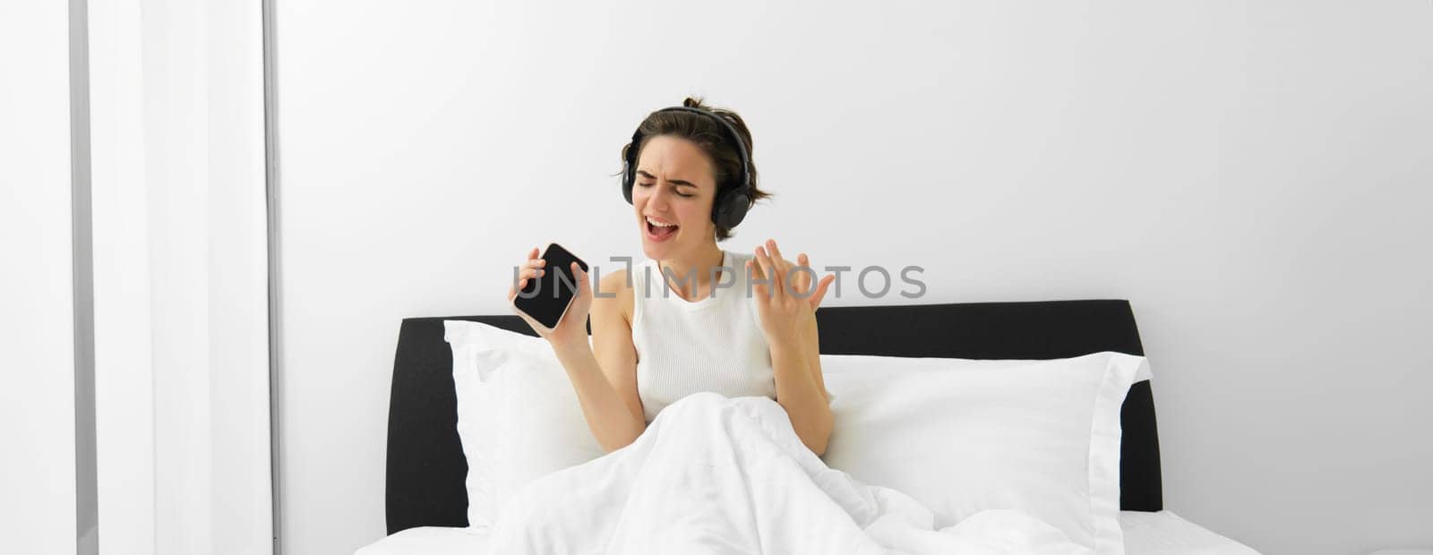 Carefree woman in bed, wakes up and listens to music in wireless headphones, dancing and singing, using mobile phone as microphone.