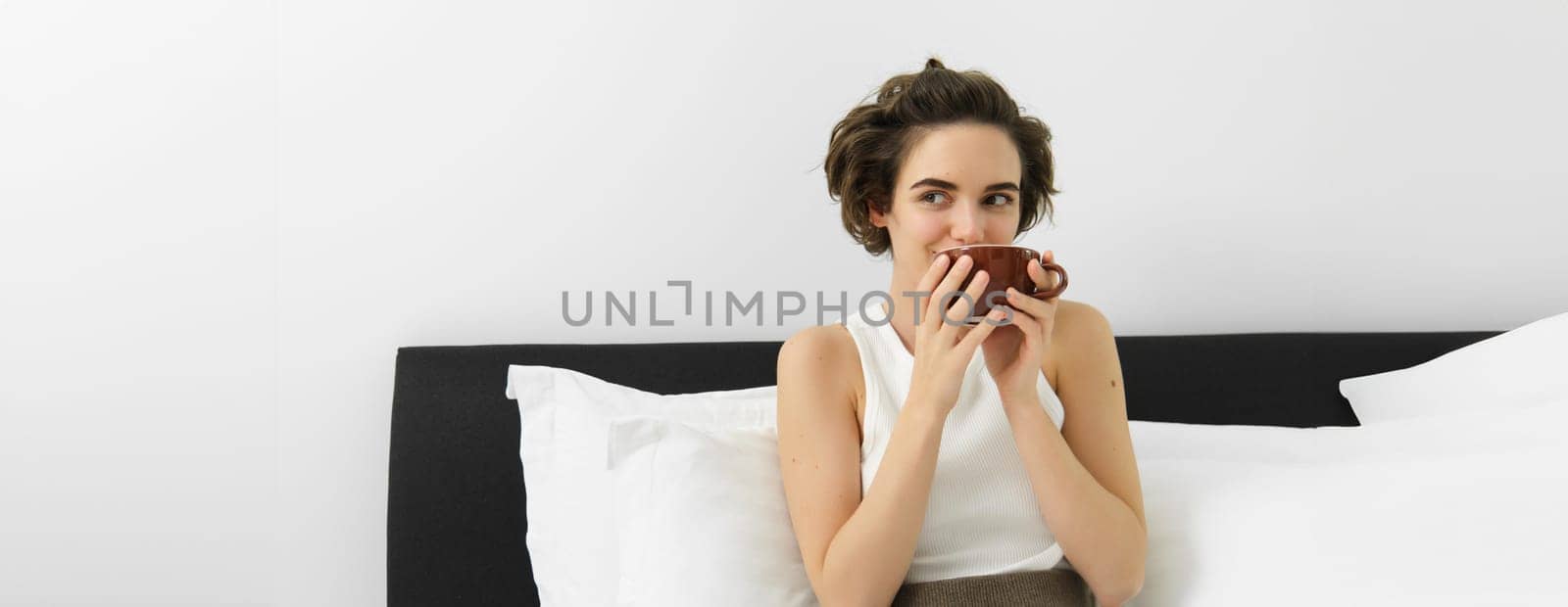 Portrait of beautiful young woman resting in her bedroom, lying in bed and drinking coffee, having her morning cup and smiling.