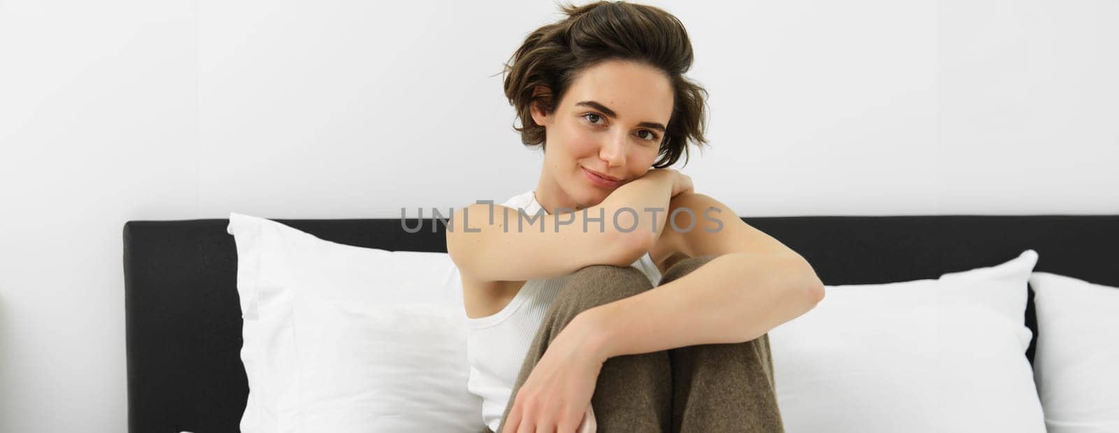 Young tender woman in her cosy pyjamas, sitting on bed in bedroom, smiling with sensual smile.