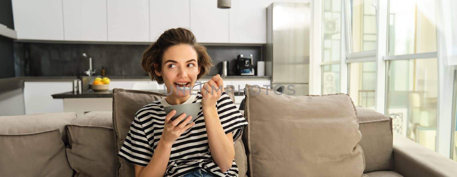 Portrait of cute young woman eating breakfast, having cereals with milk on sofa in living room, smiling and looking satisfied.