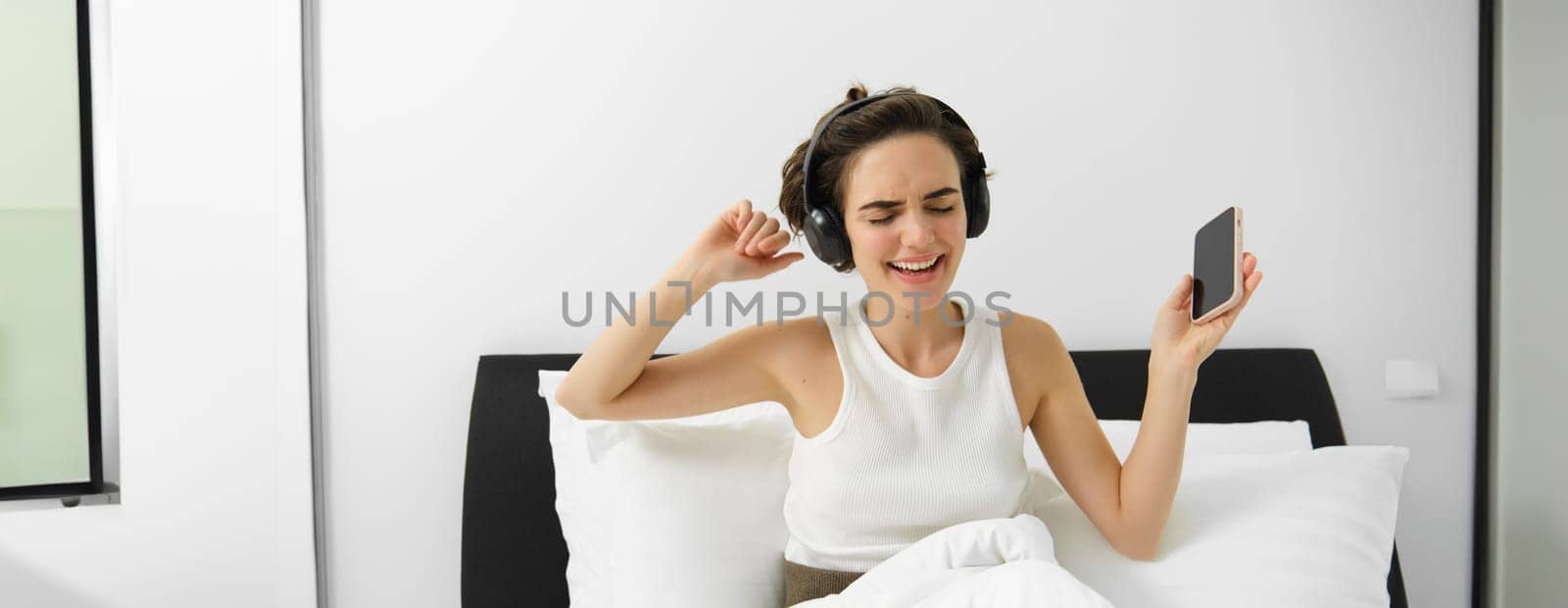 Portrait of happy young woman in bed, listening to music in wireless headphones, dancing in her bedroom, holding mobile phone in hand. Copy space