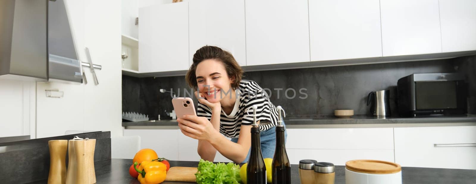 Portrait of smiling young woman cooking in the kitchen, watching videos on smartphone app, scrolling social media, looking for healthy recipes from vegetables, posing at home.