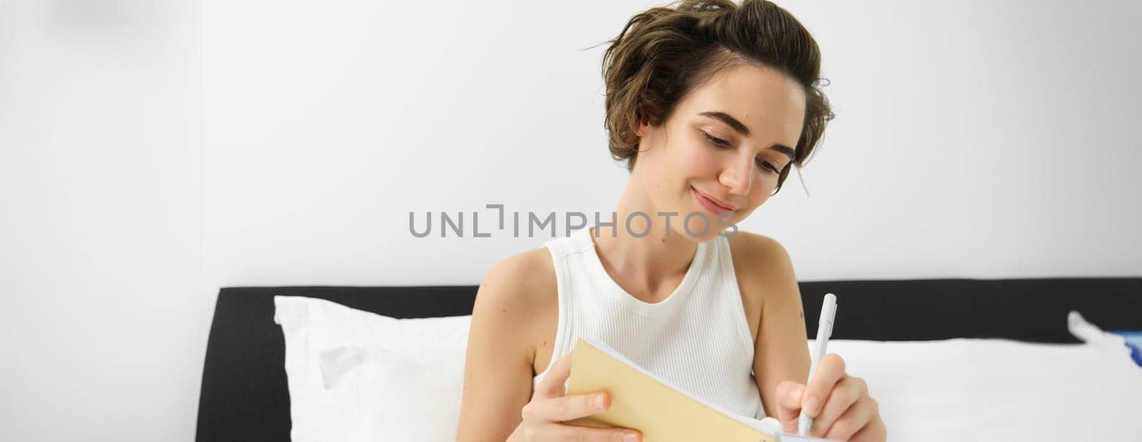 Portrait of young woman in bed, working or studying, writing in her journal, reading notebook notes, sharing thoughts with diary and smiling.