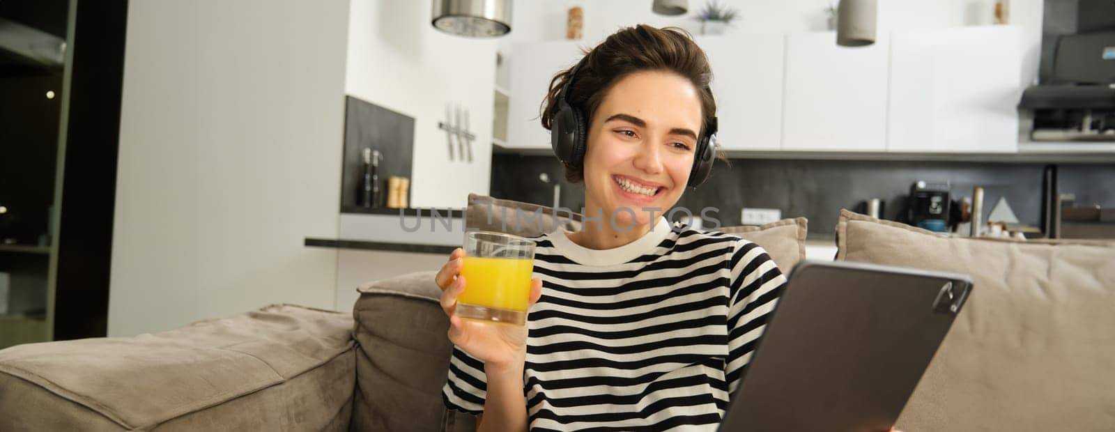 Close up portrait of young brunette woman in living room, watching funny video or tv series on digital tablet, drinking orange juice, sitting on sofa and laughing.