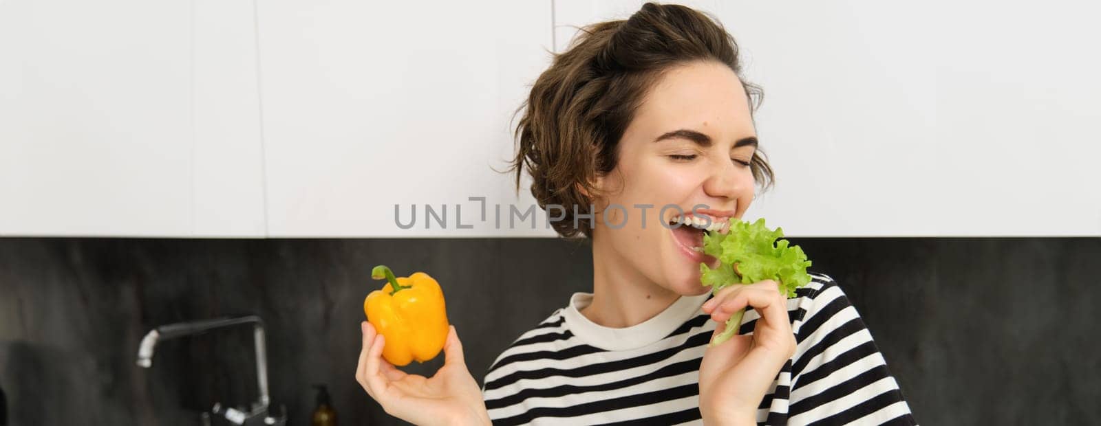 Portrait of carefree vegetarian girl, eating vegies, bite lettuce leaf with happy smiling face, having healthy snack, likes vegetables, stands in the kitchen.