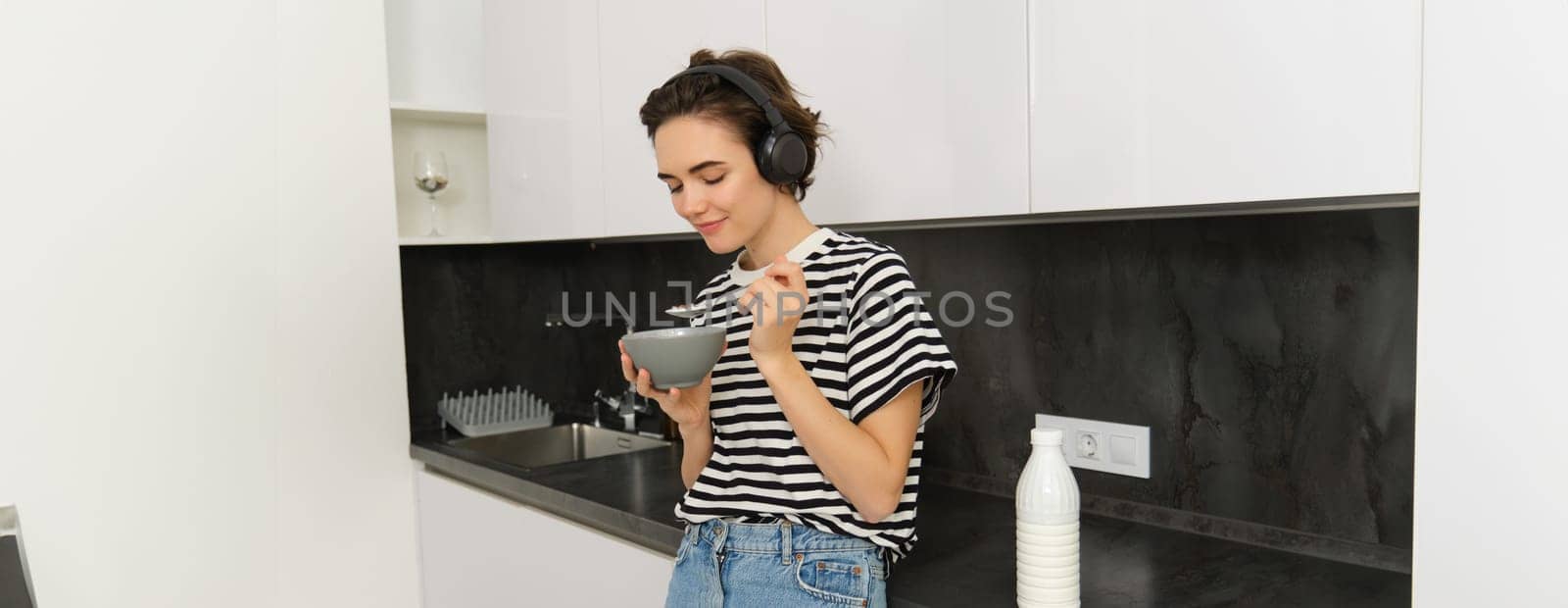 Candid young woman eats breakfast, cereals from bowl, stands near kitchen counter, listens music in wireless headphones.