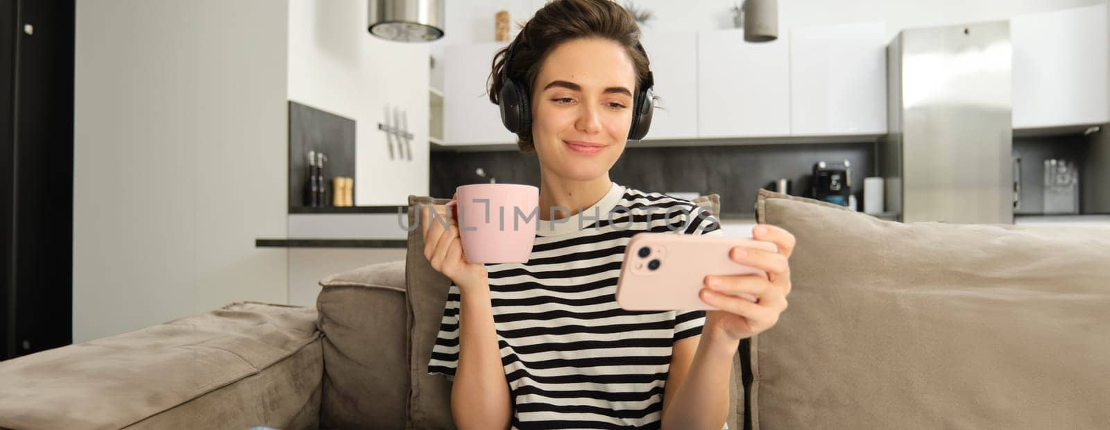 Portrait of woman in her living room, sitting on sofa and watching videos on mobile phone, wearing headphones, drinking tea or coffee in pink cup.