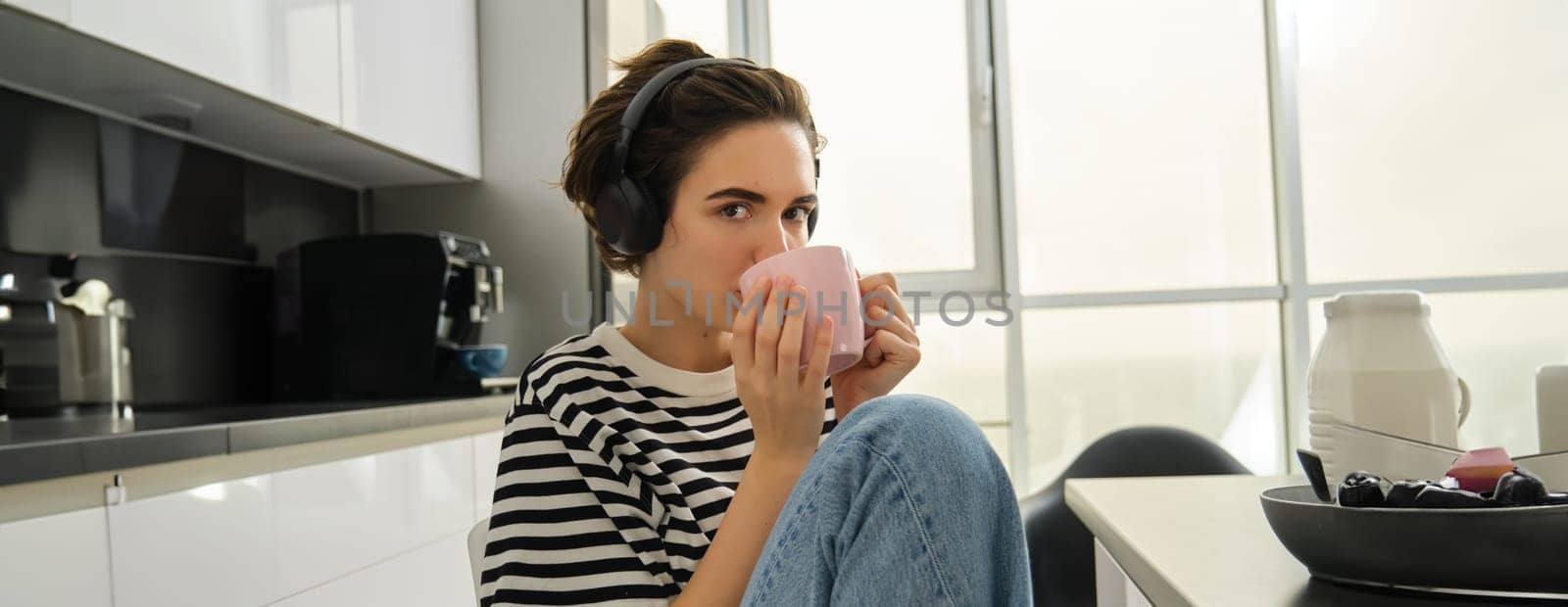 Close up portrait of smiling brunette woman, student drinks her tea and listens music or ebook in headphones, sits in kitchen and relaxes.