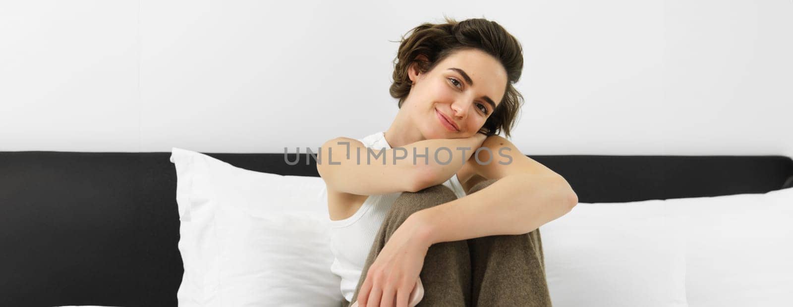 Close up portrait of young female model in white tank-top, sitting on bed in her bedroom and smiling at camera, leaning on pillows.