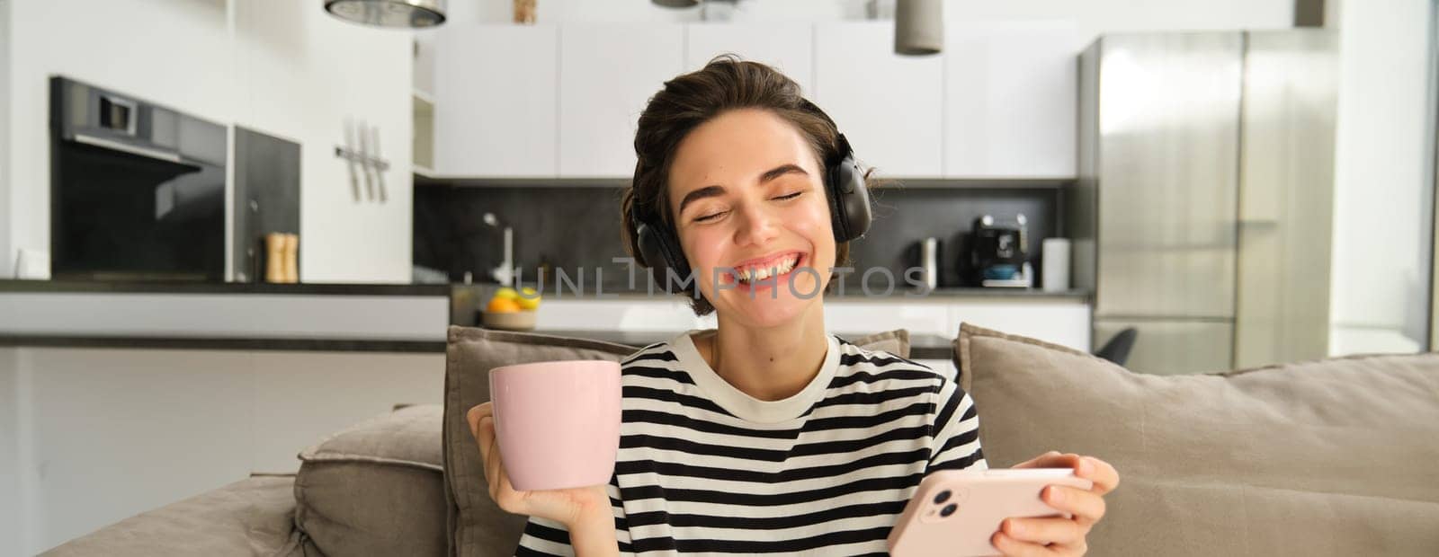 Carefree female model in headphones, sitting on sofa with smartphone and cup of tea, watching videos in wireless earphones on mobile phone app, resting at home by Benzoix