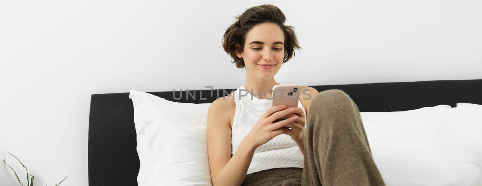 Portrait of young beautiful woman lying in bed, resting in bedroom, messaging, using mobile phone, holding smartphone and smiling, scrolling social media, spending comfort time at home by Benzoix
