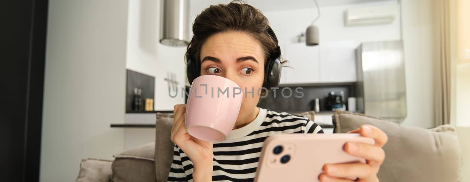Portrait of girl sipping tea and watching tv show on smartphone with interest, stating at mobile phone amazed, sitting on sofa in living room.