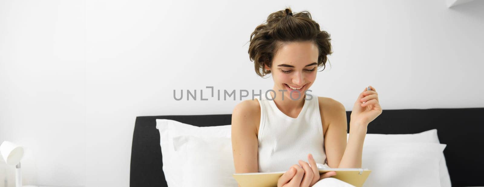 Portrait of woman sitting in bed with journal, writing in diary with pen, reading notebook, studying or doing homework from her bedroom. Copy space