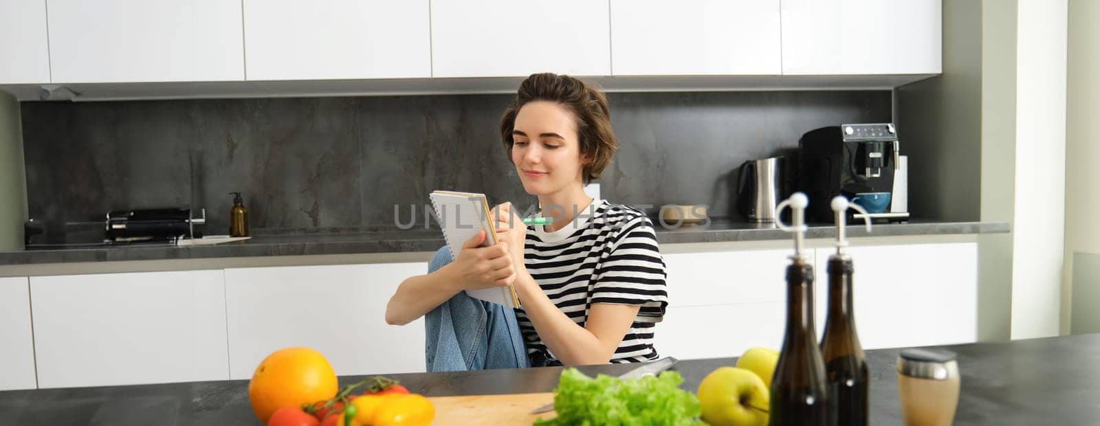 Portrait of beautiful modern woman, girl cooking with vegetables, holding notebook, writing down recipe, making notes, dinner plans, creating meal list for week, sitting in kitchen.