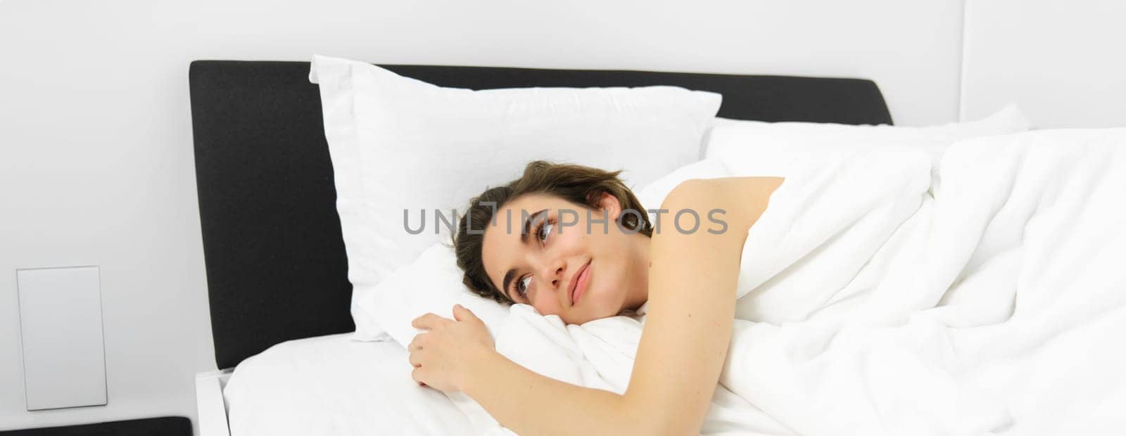 Portrait of young woman relaxing, lying in bed on soft white pillows, covered in linen comfortable sheets, smiling and looking outside with dreamy smile.