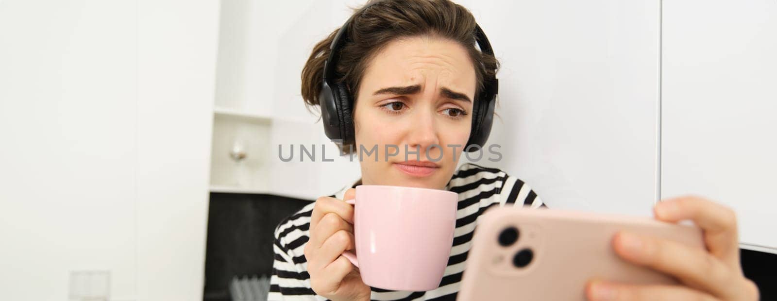 Portrait of woman with sad face, watching something on mobile phone, social media app, drinking tea, wearing wireless headphones.