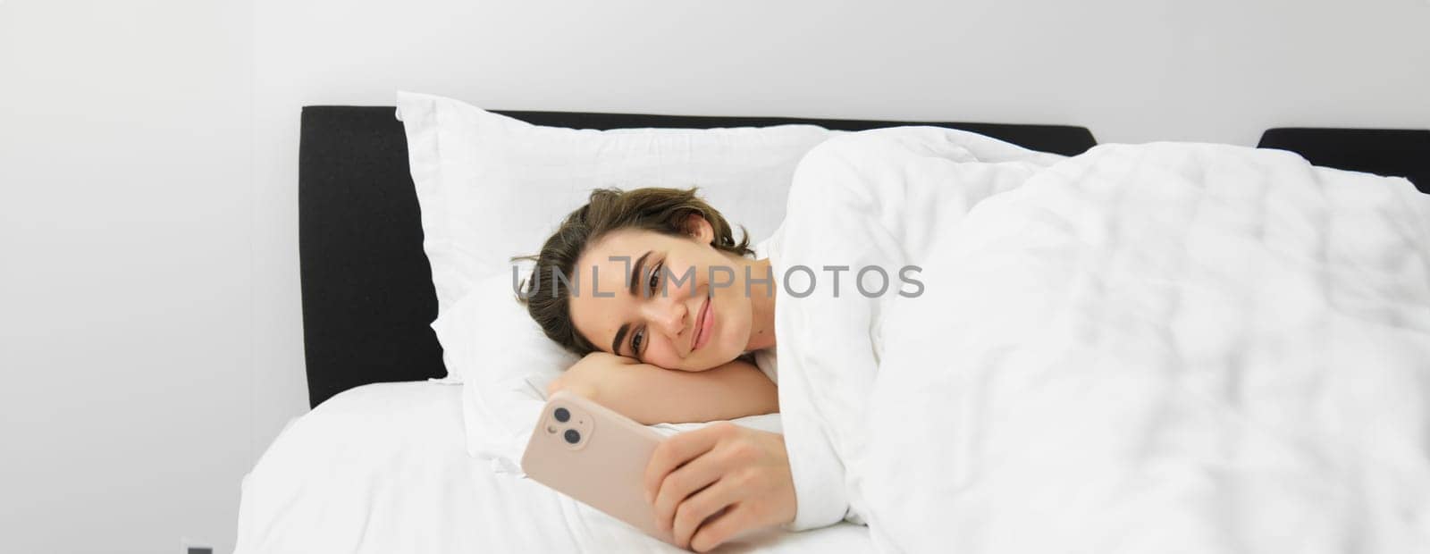 Smiling beautiful woman, lying in bed with smartphone, looking at mobile phone, using application, reading message and looking pleased.