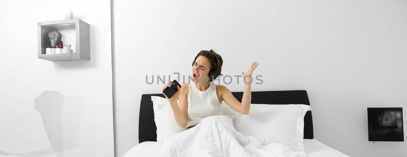 Image of modern, beautiful woman sitting on bed, singing into smartphone, listening music in wireless headphones by Benzoix