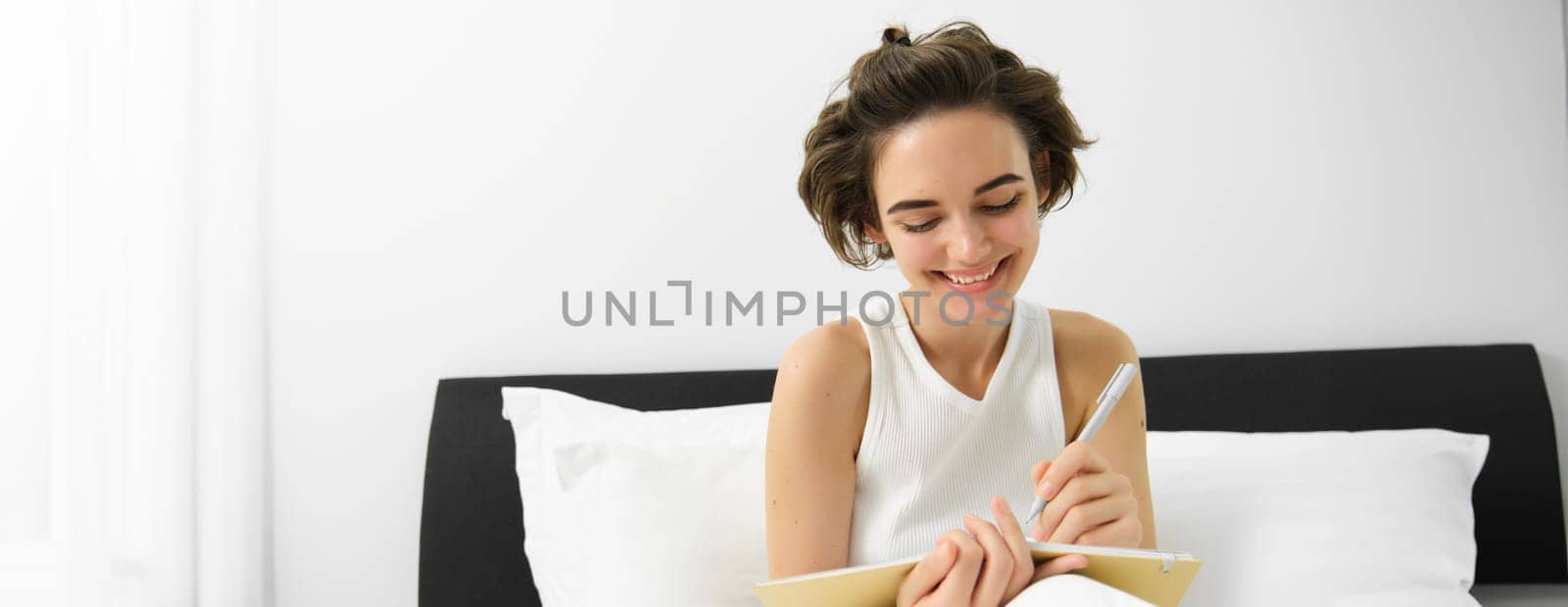 Portrait of smiling, happy young woman sitting in bed, writing in journal, making notes in diary, doing homework in bedroom by Benzoix