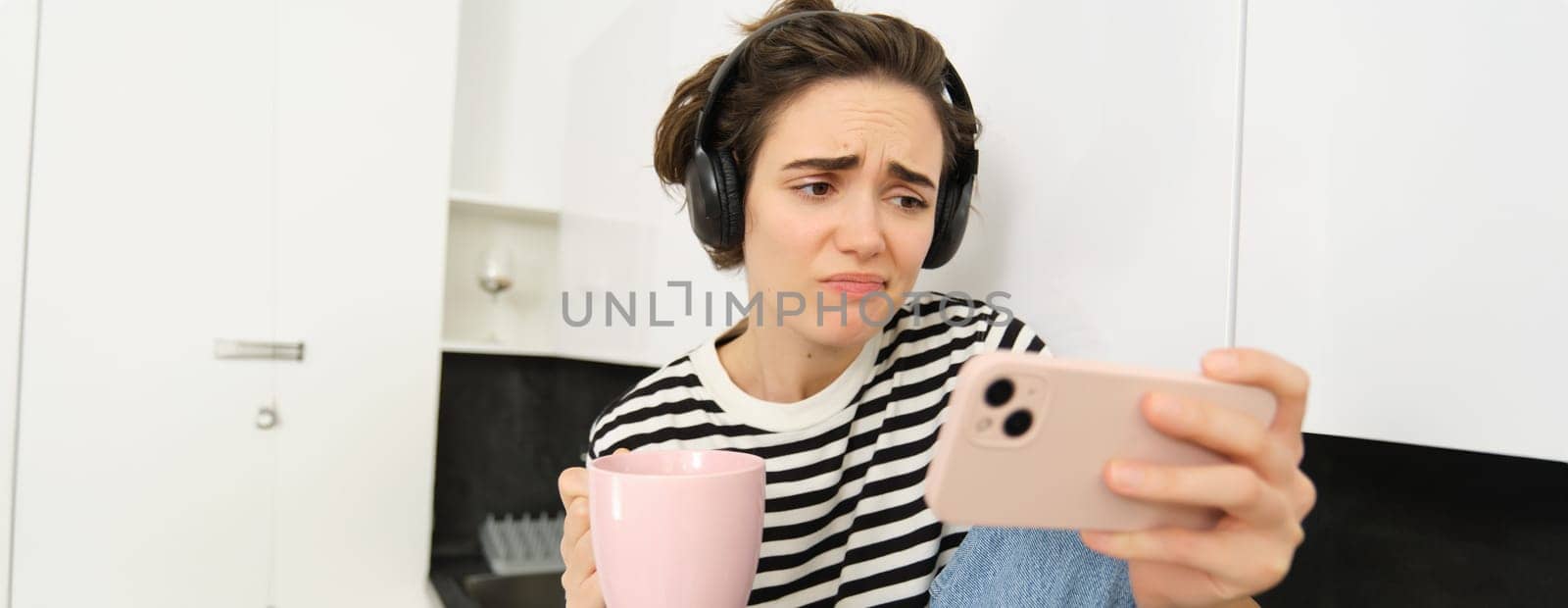 Portrait of woman looking with sympathy and sad face at smartphone screen, watching video on mobile phone app, drinking tea or coffee in the kitchen, frowning and looking with pity.