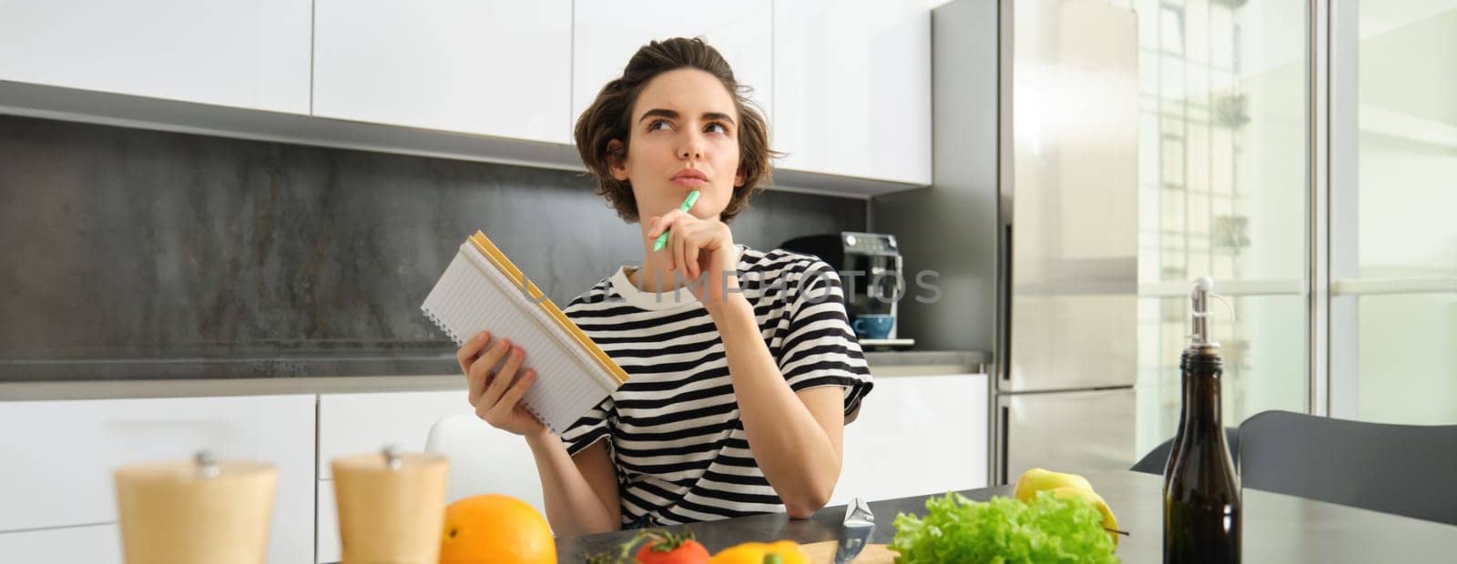 Portrait of thinking woman with notebook, cooking, writing down recipe ingredients, deciding on a meal for dinner, sitting near vegetables and chopping board in kitchen.