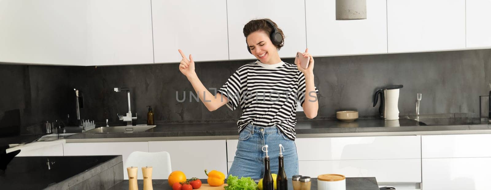 Dancing girl cooking salad in the kitchen, listening music in wireless headphones, holding smartphone in hand, making a meal, cooking at home.