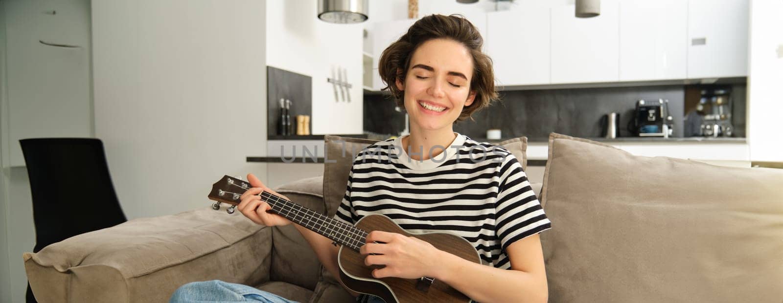 Young happy woman sitting on sofa and playing ukulele, singing and enjoying learning new musical instrument.