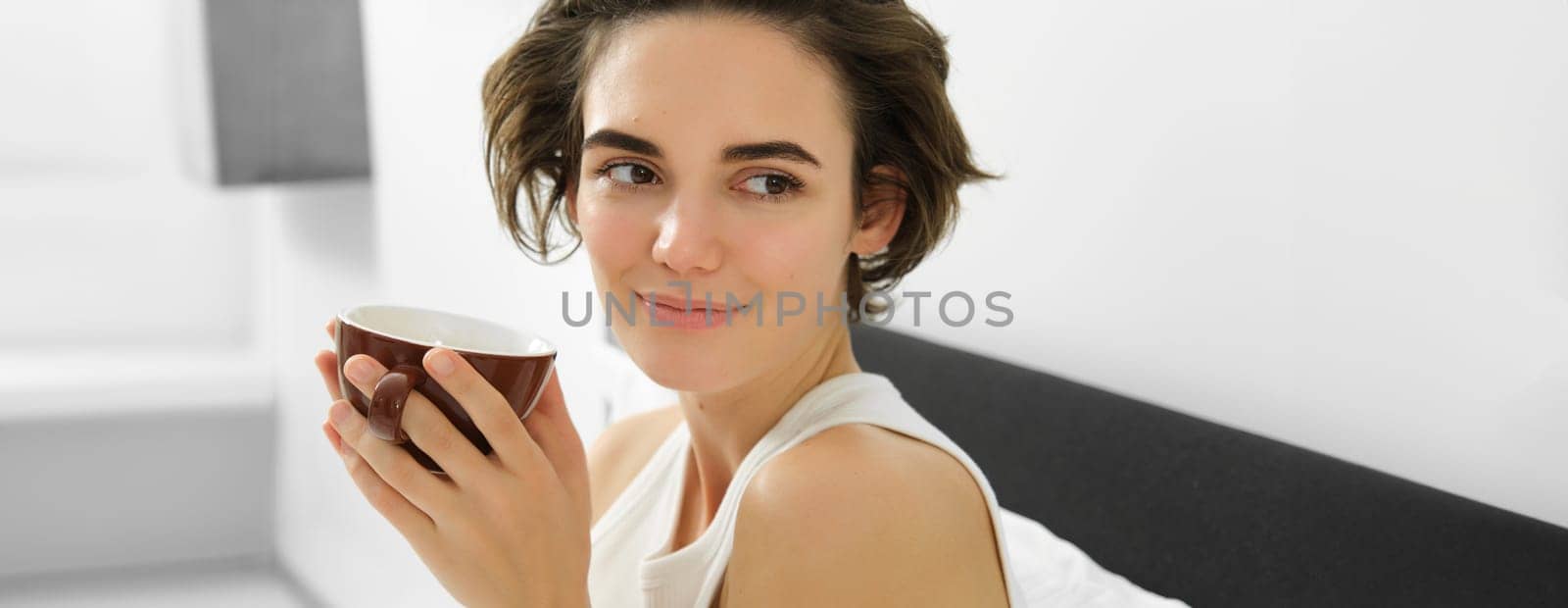 Close up of sensual young woman, looking aside and smiling, lying in bed, drinking morning cup of coffee, brown mug with drink.