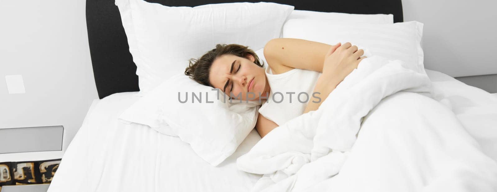 Portrait of woman feeling unwell, lying in bed and touching her belly, frowning from painful menstrual cramps, having period pain, resting in bedroom by Benzoix